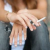 A teenager with white nail polish on their fingers, sits on a step and holds a cigarette