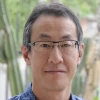 Man with dark hair, glasses and blue shirt stands outside in a wooded area