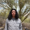 Man with long black hair and wearing a gray plaid shirt stands in front of a tree outside