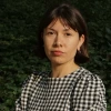 A woman with dark brown hair and wearing a black and white checked dress, stands outside with arms folded