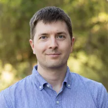 A man with short brown hair and a blue shirt stands outside in front of green foliage