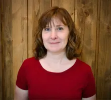 Woman with light brown hair and a red shirt stands in front of a wood paneling wall 