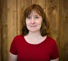 Woman with medium length light brown hair and wearing a red shirt smiles while in front of a wood paneled wall