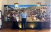 Man wearing a plaid shirt, dark pants, and glasses stand in front of a mural showing the past of Santa Cruz County