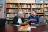 A professor and student sit together at a table in front of bookshelves