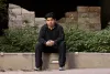 Young man with black hair and wearing all black, sits outside on a low concrete wall